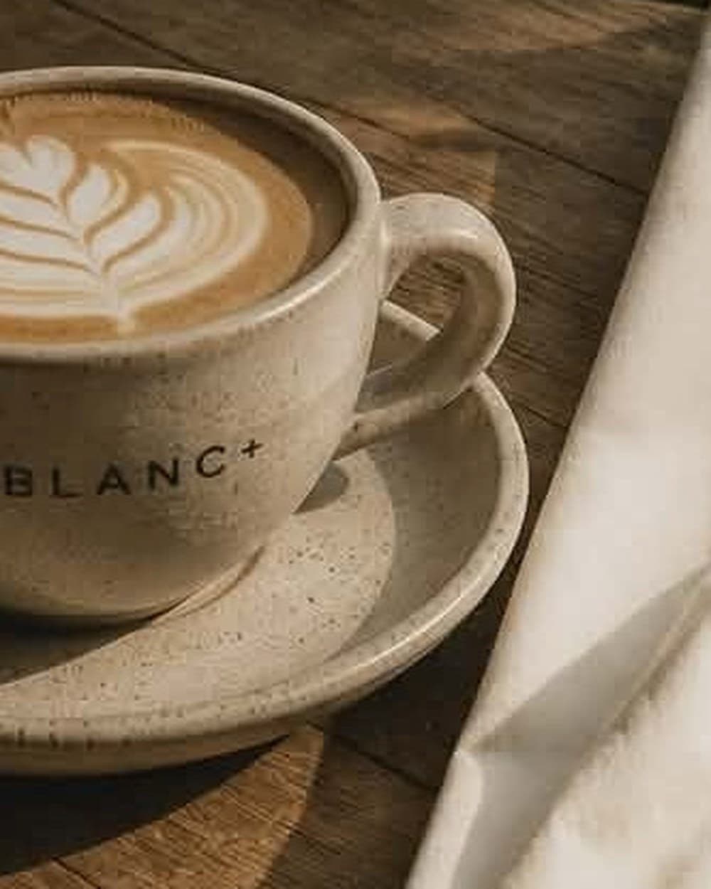 A Blanc+ branded ceramic cup of coffee on a wooden table beside a folded cream tee